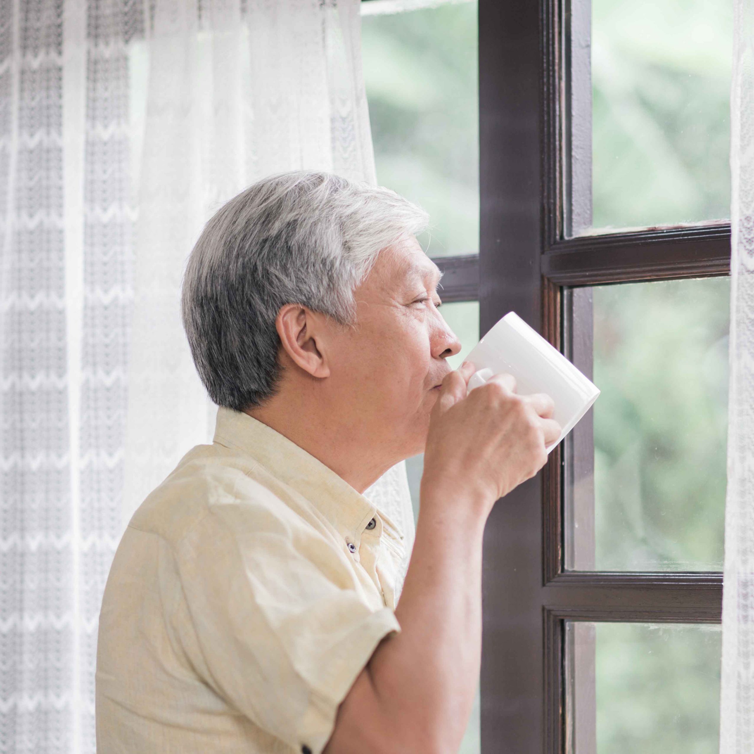Happy Asian elderly man smiling and drinking a cup of coffee or tea near the window in living room, senior asia male open the curtains and relax in the morning. Lifestyle senior men at home concept.